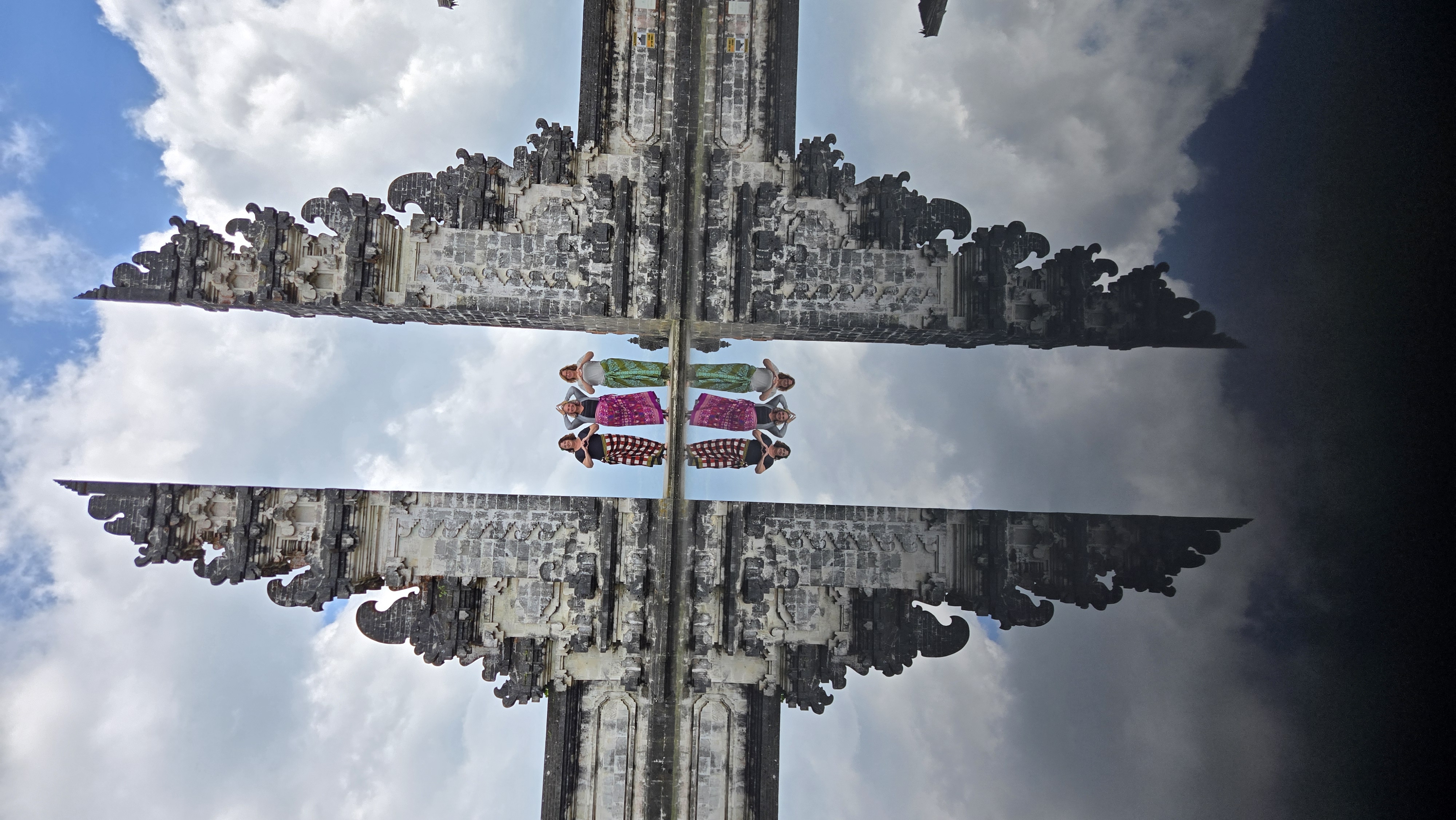 Three women stand between two carved stone temple gates reflected in water, wearing colorful traditional sarongs