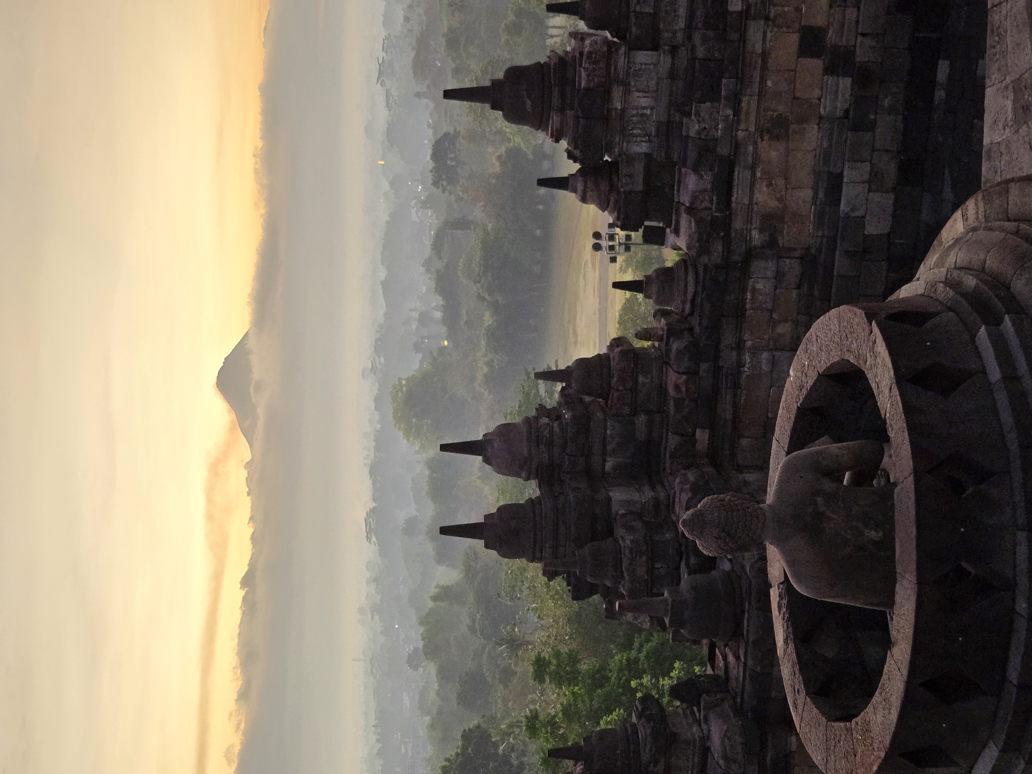 A stone Buddha statue in the foreground with tiered temple structures and mountains visible behind in soft morning light