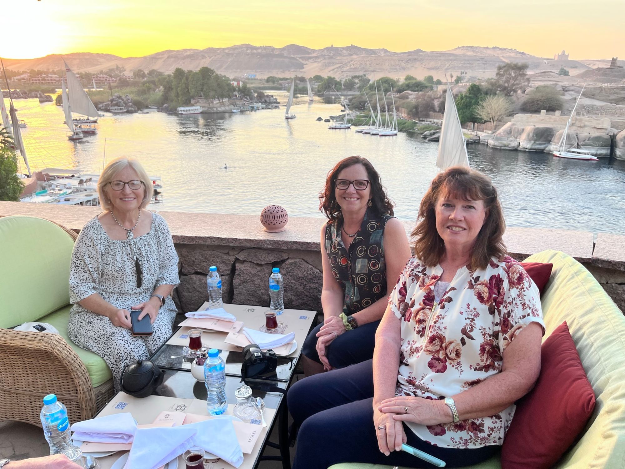Three women seated outdoors overlooking the Nile River at sunset with sailboats in the background