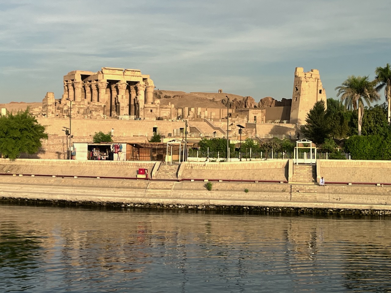 Ancient Egyptian temple ruins viewed from across the Nile River with palm trees lining the shore