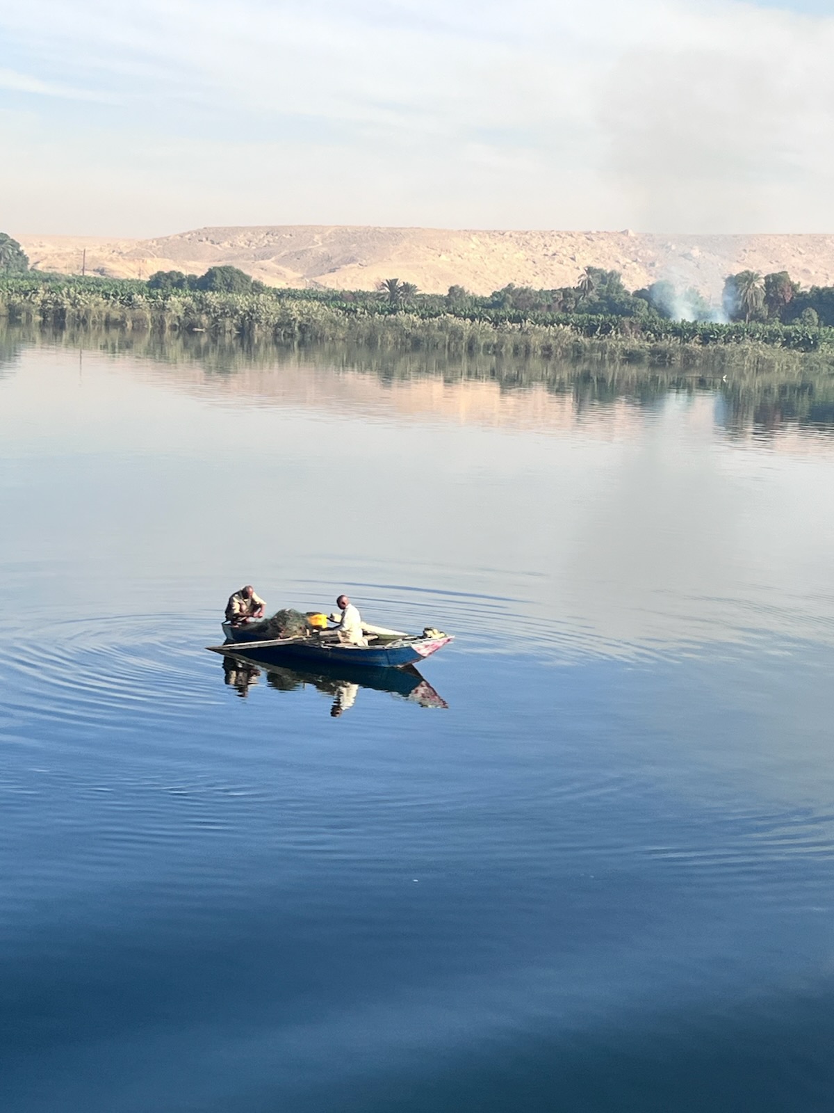 Fisherman in a small blue boat on the calm Nile River with green vegetation and desert hills beyond