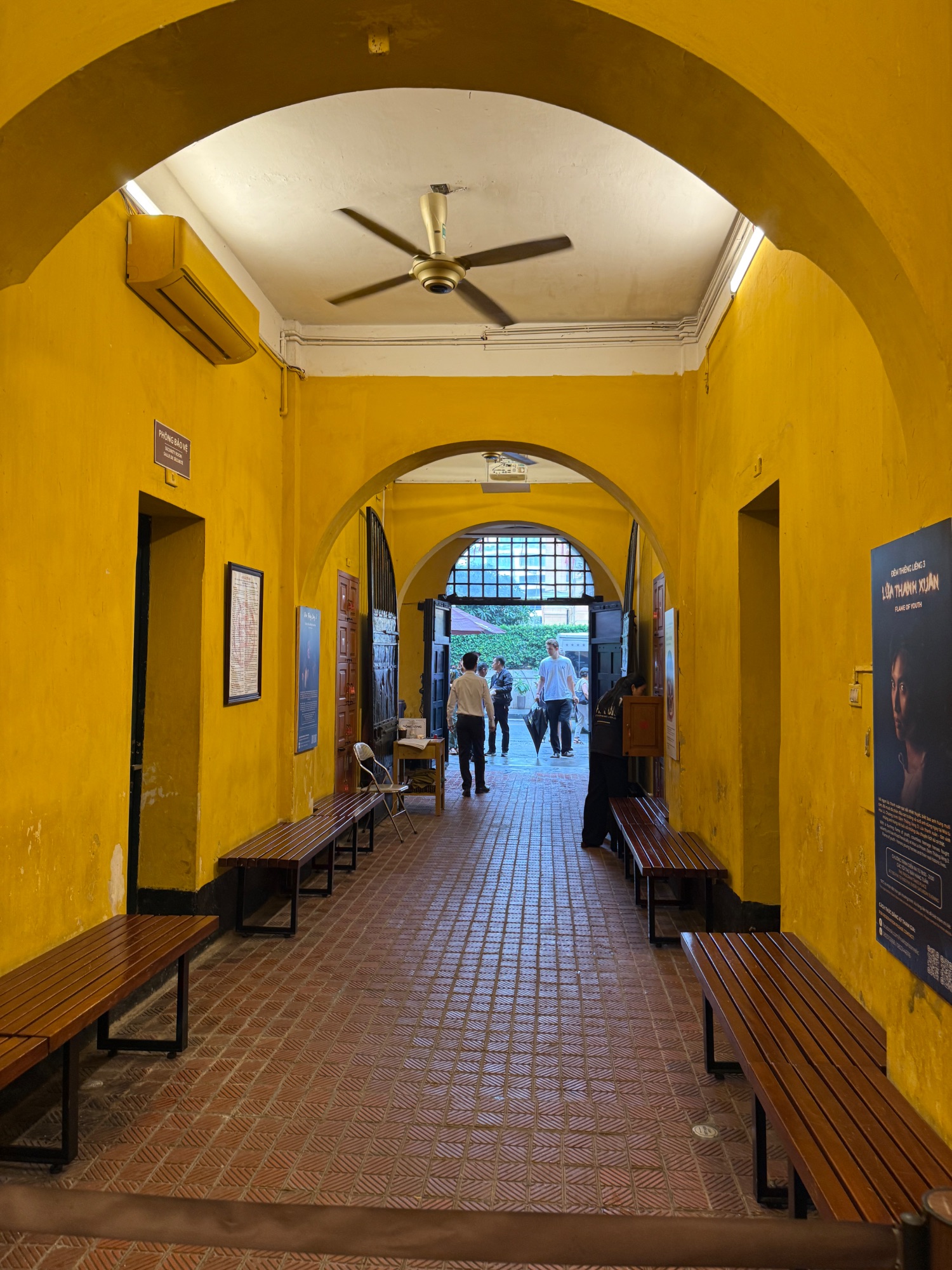 Bright yellow arched corridor with wooden benches, ceiling fan, and people walking toward daylight at the end