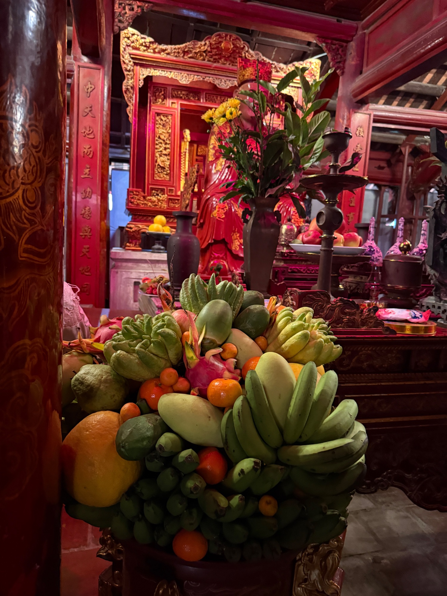Red and gold ornate altar with fresh fruit offerings, flowers in vases, and traditional Vietnamese religious decorations