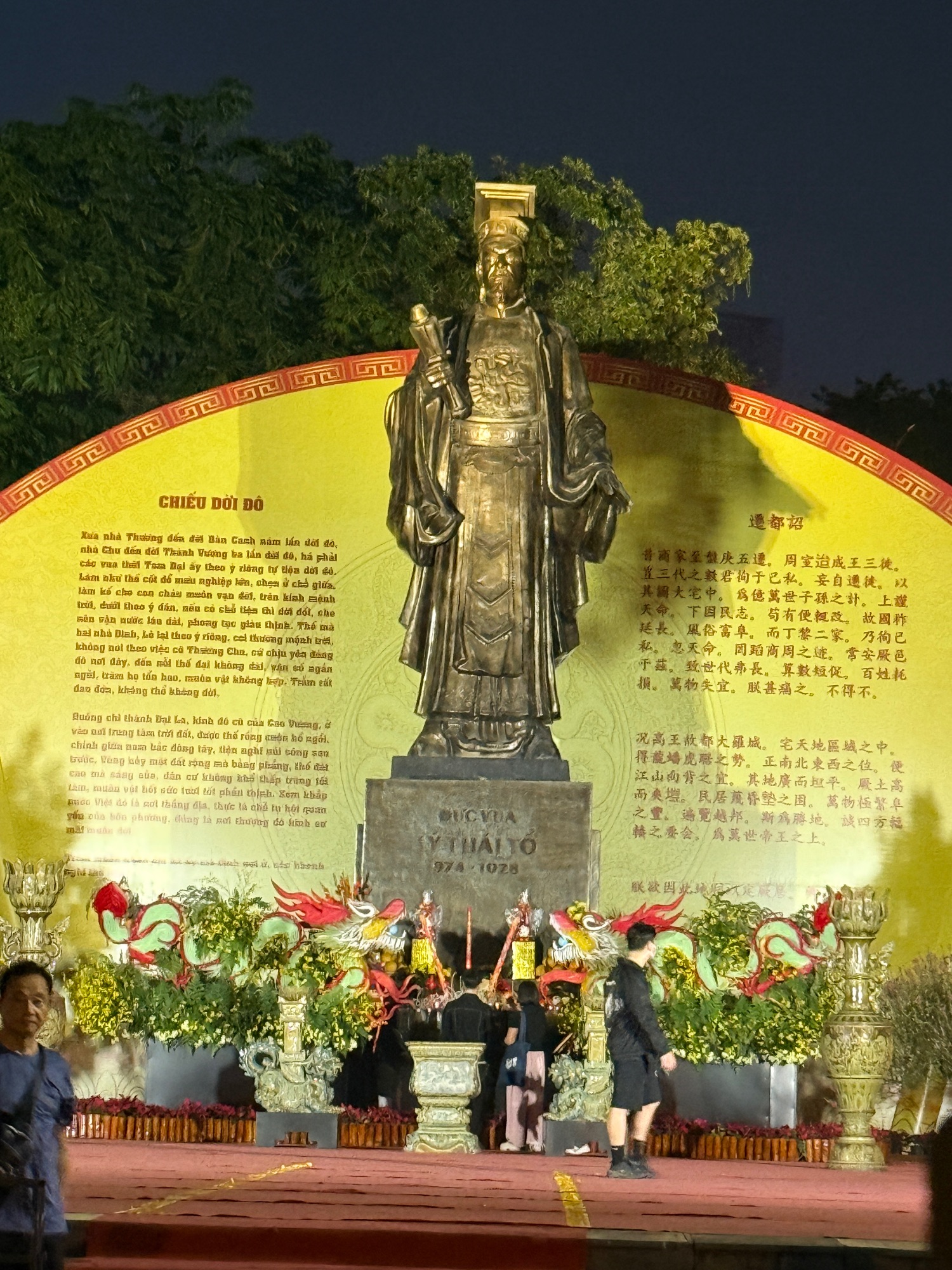 Bronze statue of a historical Vietnamese figure on illuminated yellow backdrop with ceremonial flowers and incense