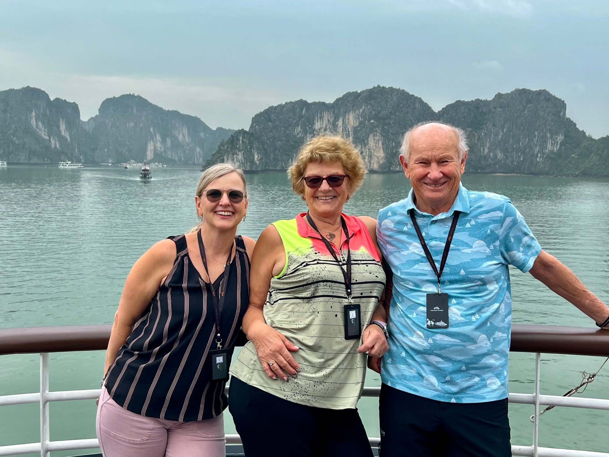 Three people on a boat railing with Ha Long Bay's dramatic limestone karsts and turquoise water in background