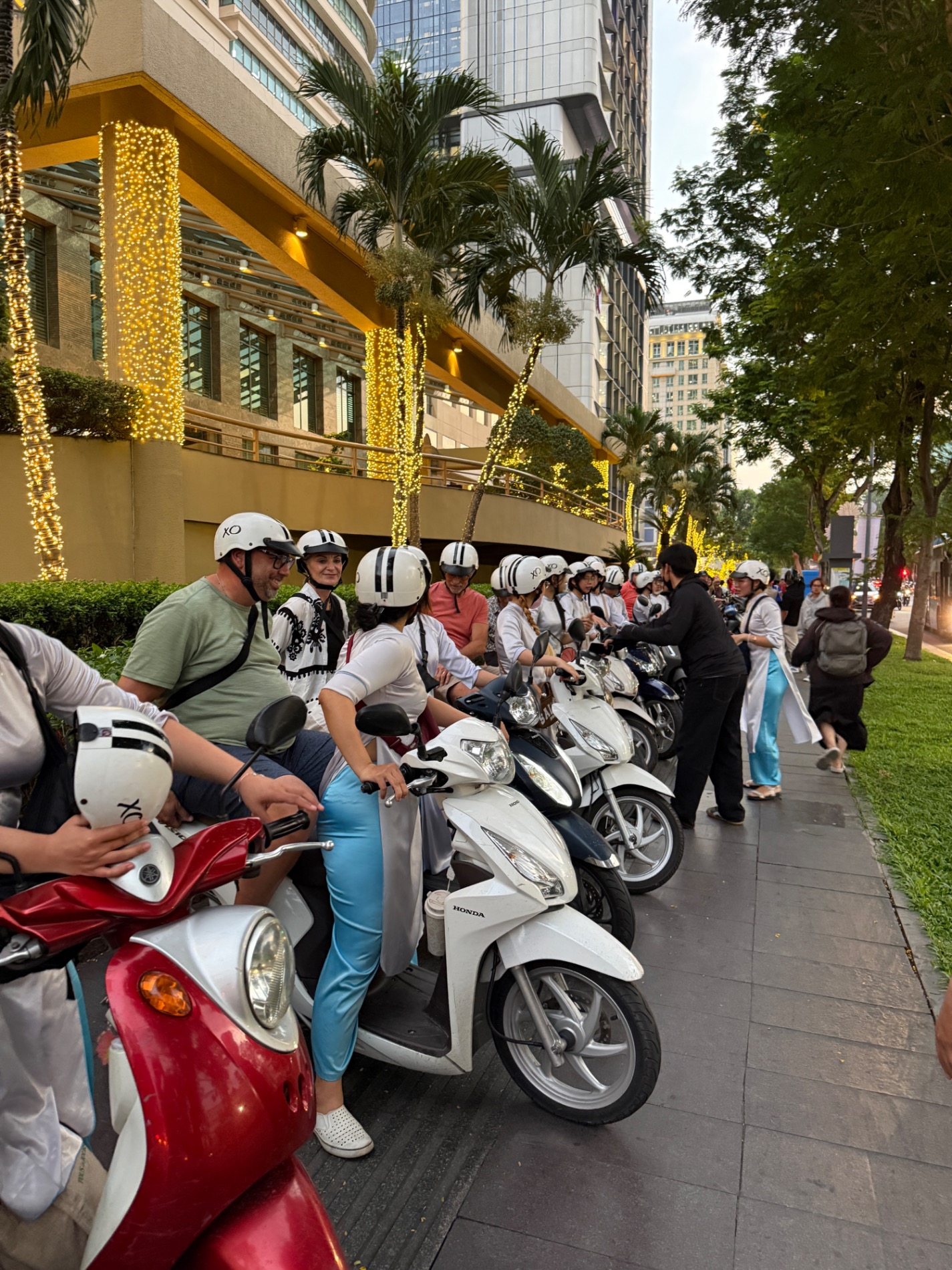 Scooter riders lined up on urban street with palm trees and modern buildings in Ho Chi Minh City