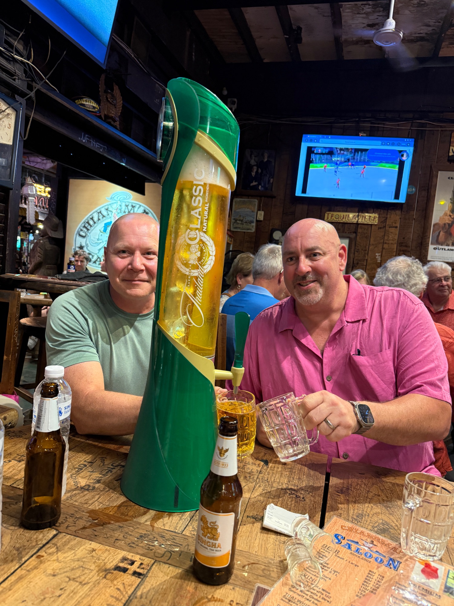 Two men at a bar counter with a large green beer tap dispenser and bottles, smiling at the camera