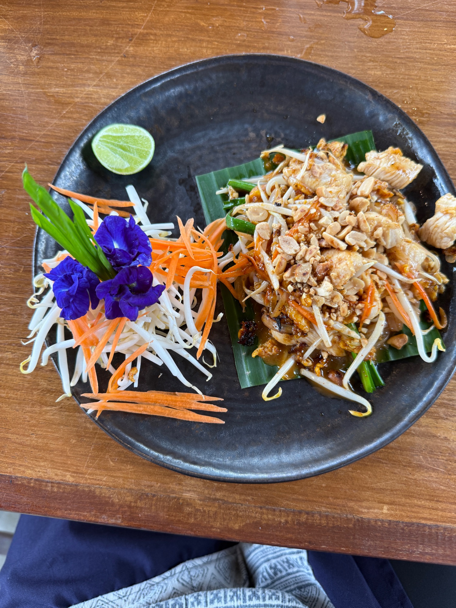 Beautifully plated Thai dish with shredded vegetables, edible purple flowers, and lime on a dark plate