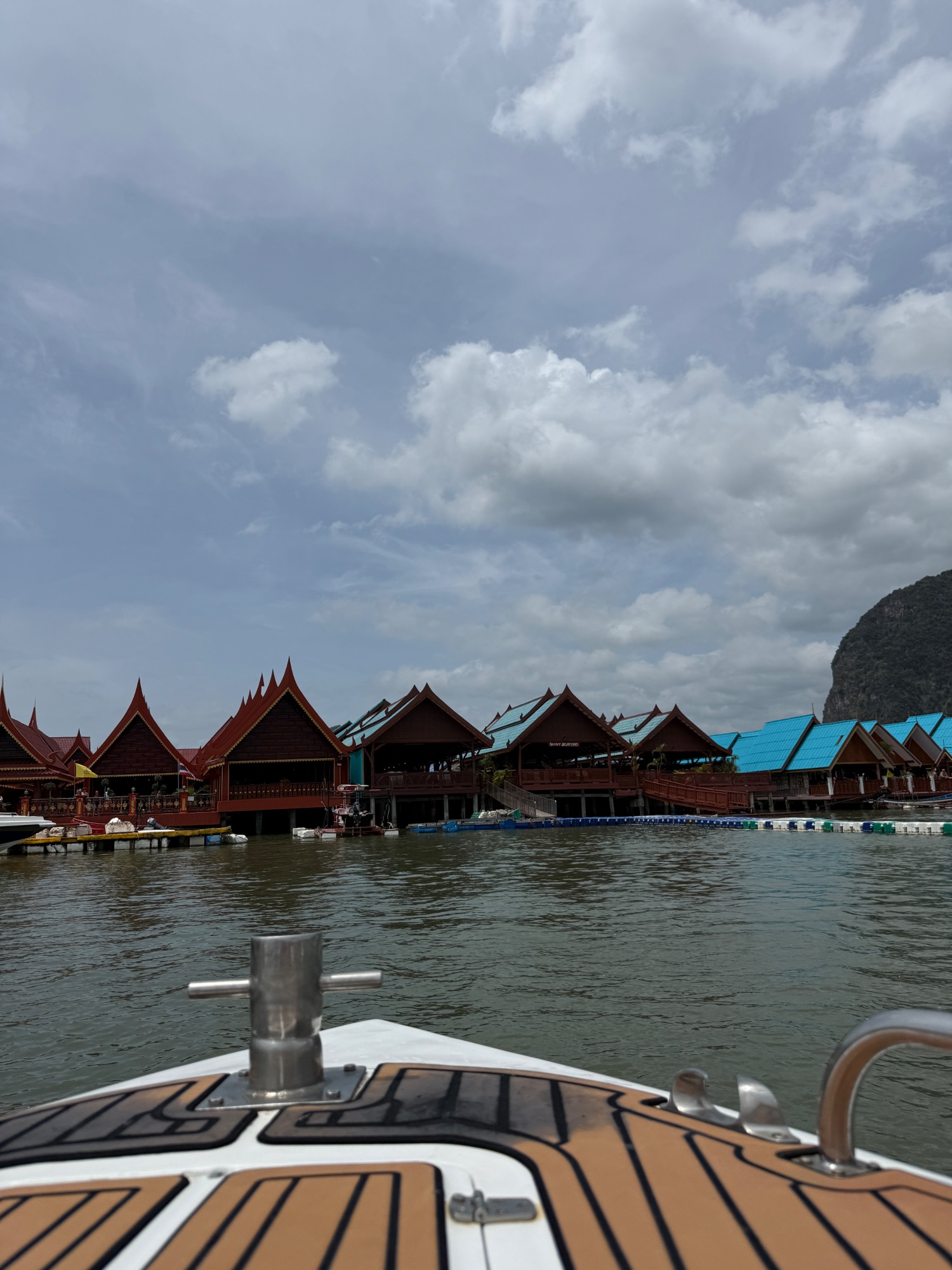 View from a boat showing traditional red-roofed wooden structures along the waterfront with limestone cliffs behind