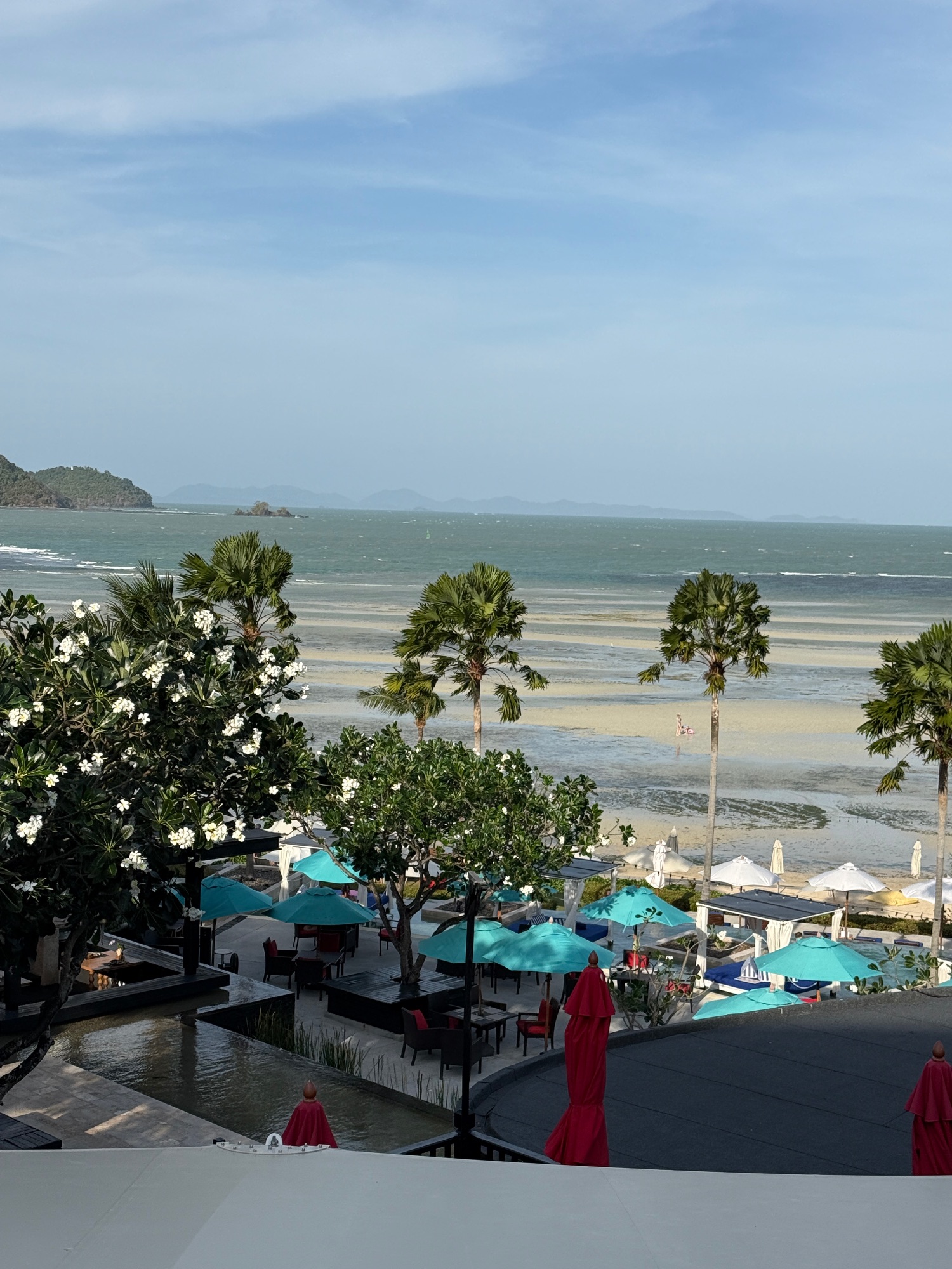 Beachfront resort with turquoise umbrellas, palm trees, and a sandy beach with distant islands visible