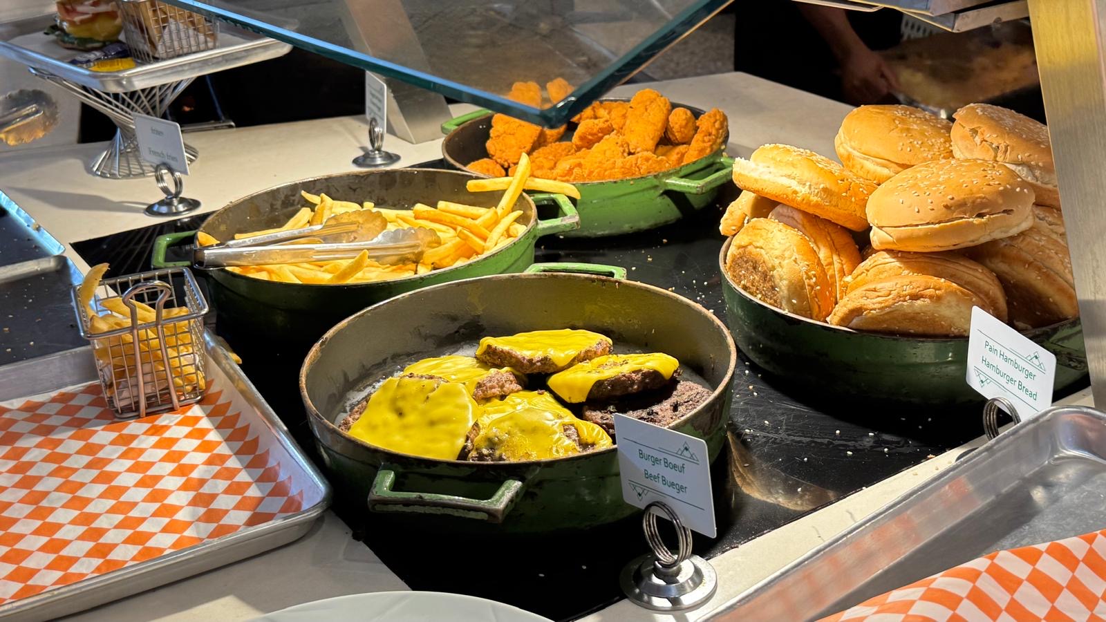 Buffet display of beef burgers with cheese, fries, and bread rolls in green serving trays