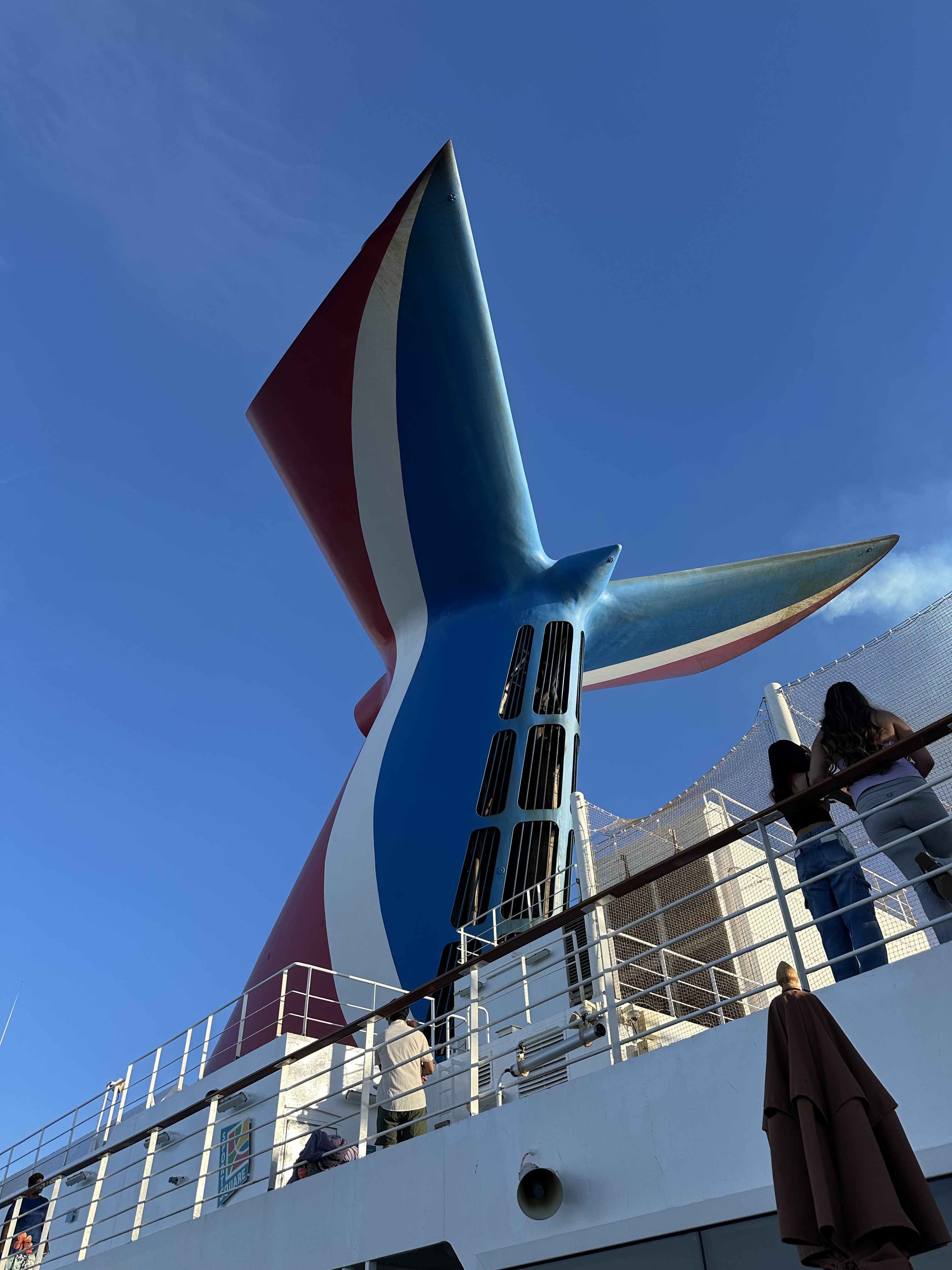 Blue and red funnel of a cruise ship with red, white and blue striped tail fin against clear sky