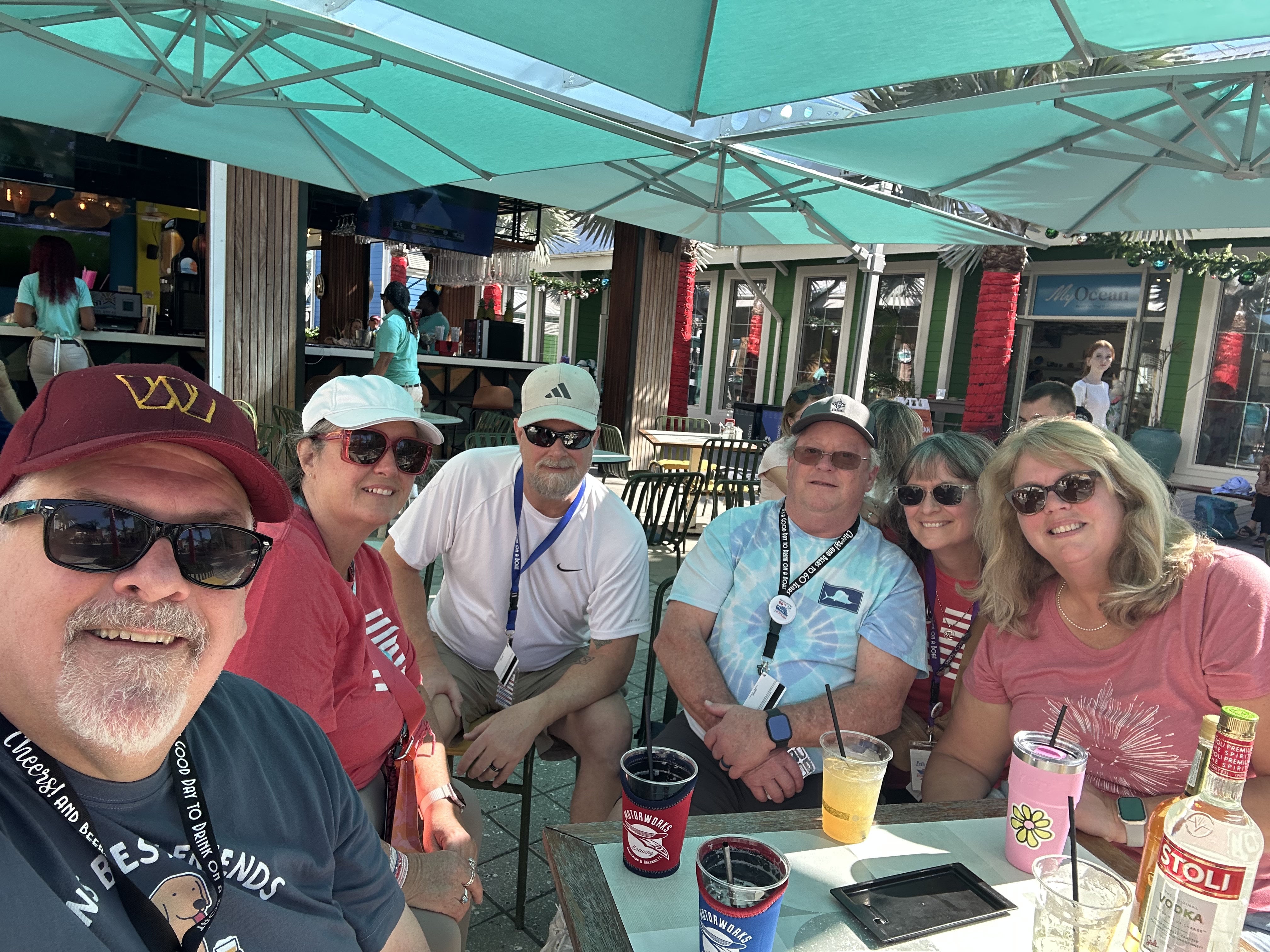Group of cruise passengers dining outdoors under turquoise umbrellas at a waterfront restaurant