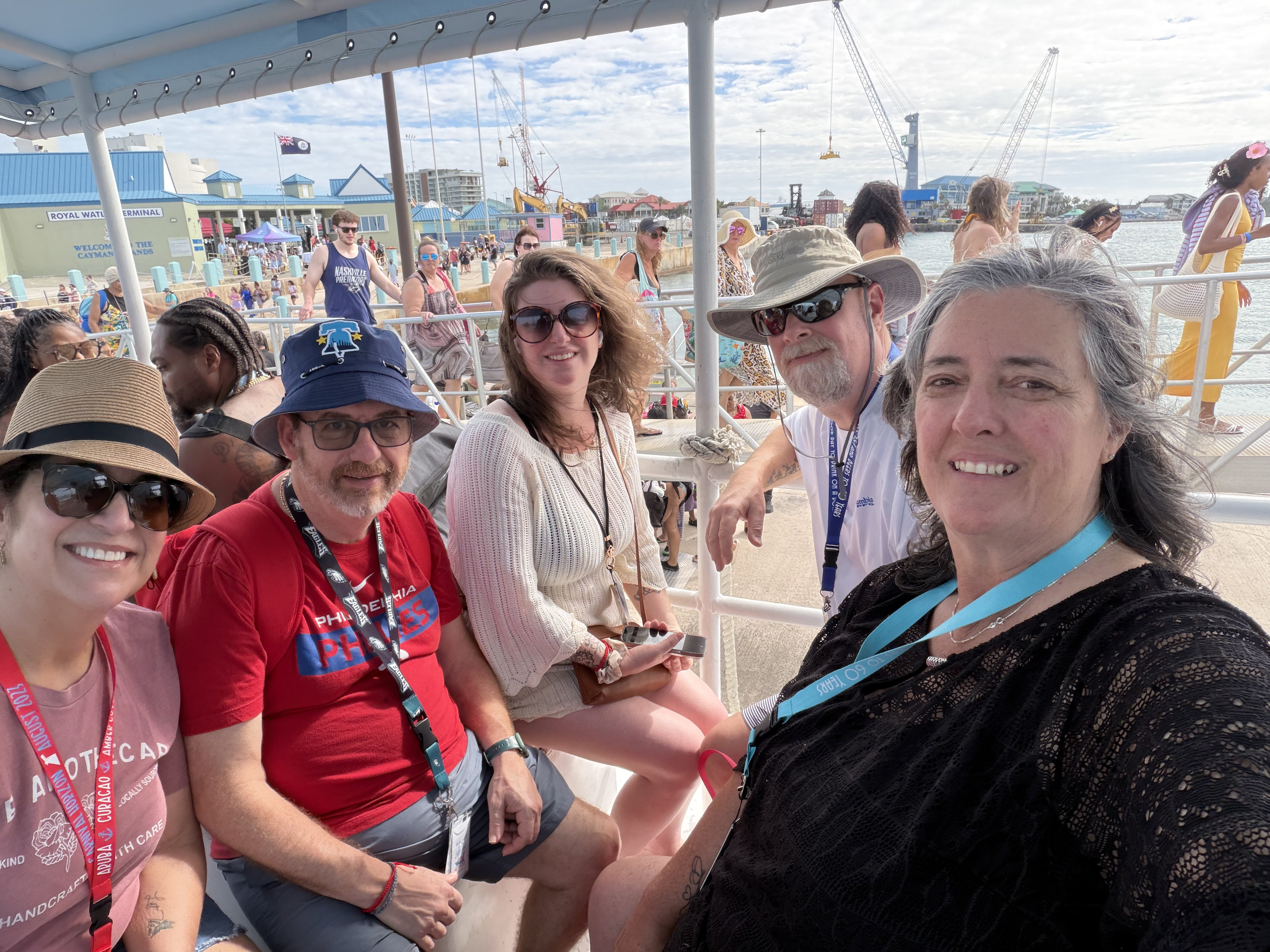 Group of five people smiling on ship's deck with waterfront and docked ships visible
