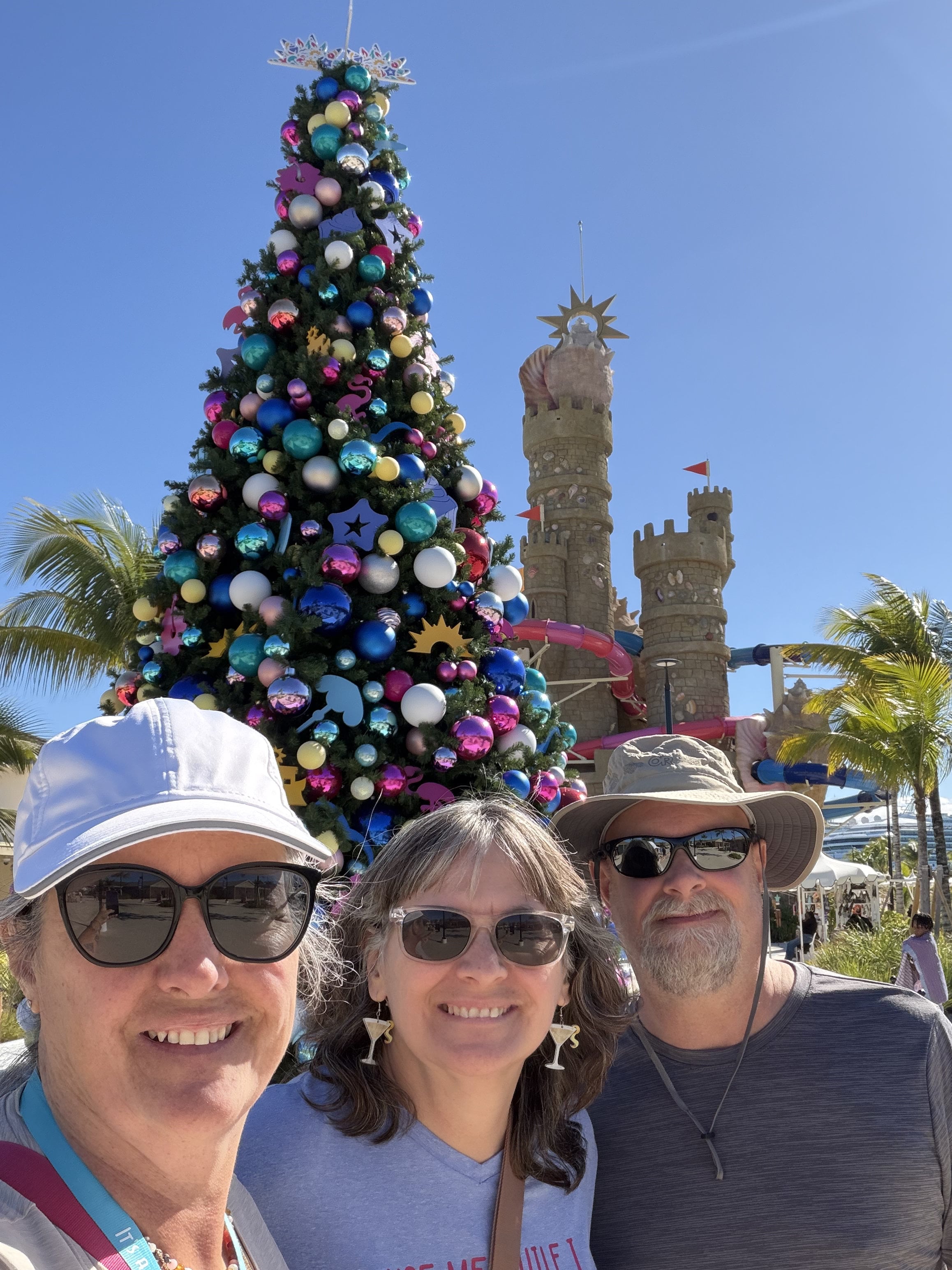 Three people posing before a colorful Christmas tree with castle towers in background at port