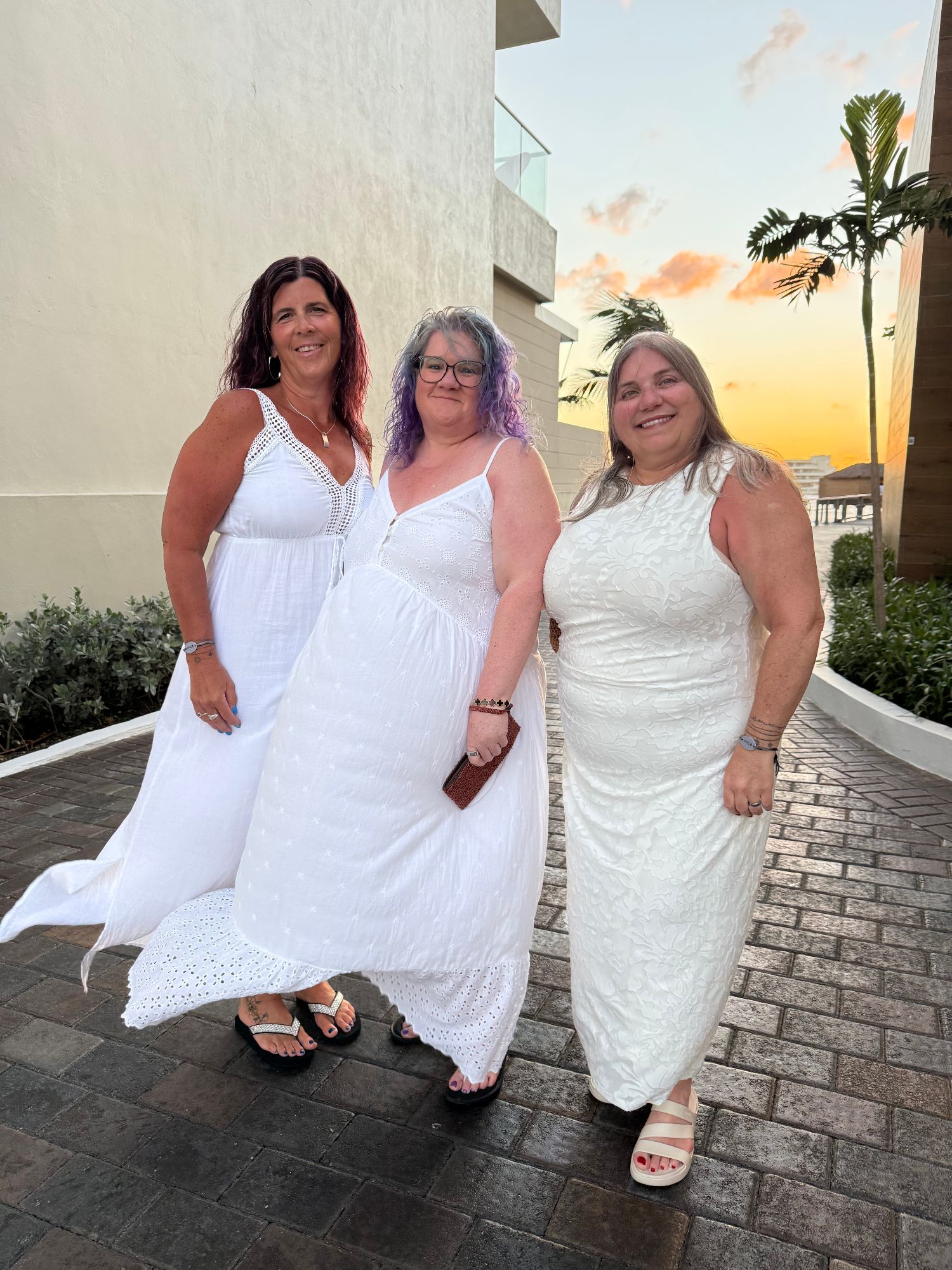 Three women in white dresses pose together at sunset on a modern terrace with palm trees and ocean views