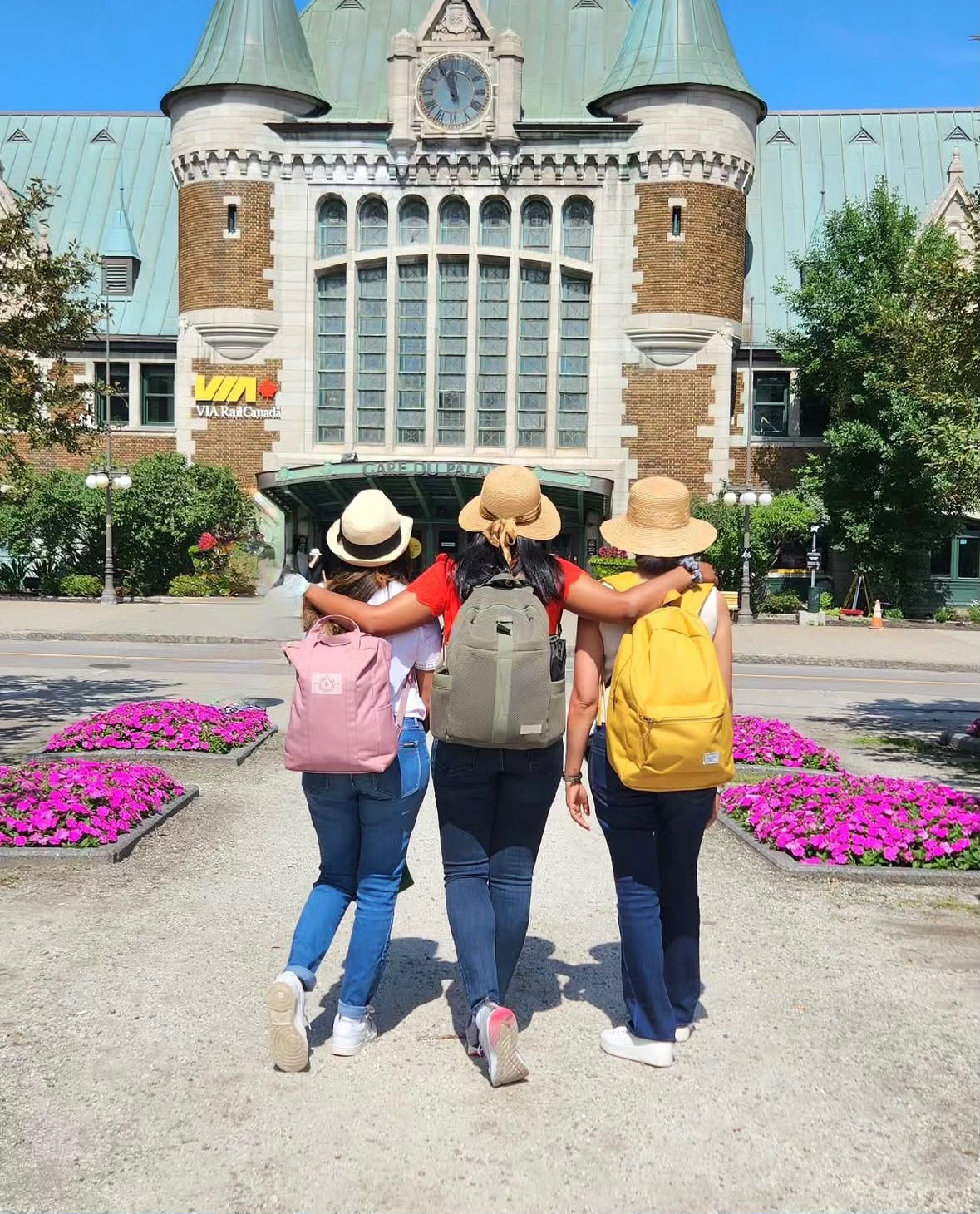 Three young women from behind wearing colorful backpacks and sun hats standing in front of a historic brick building with turquoise roof in Quebec