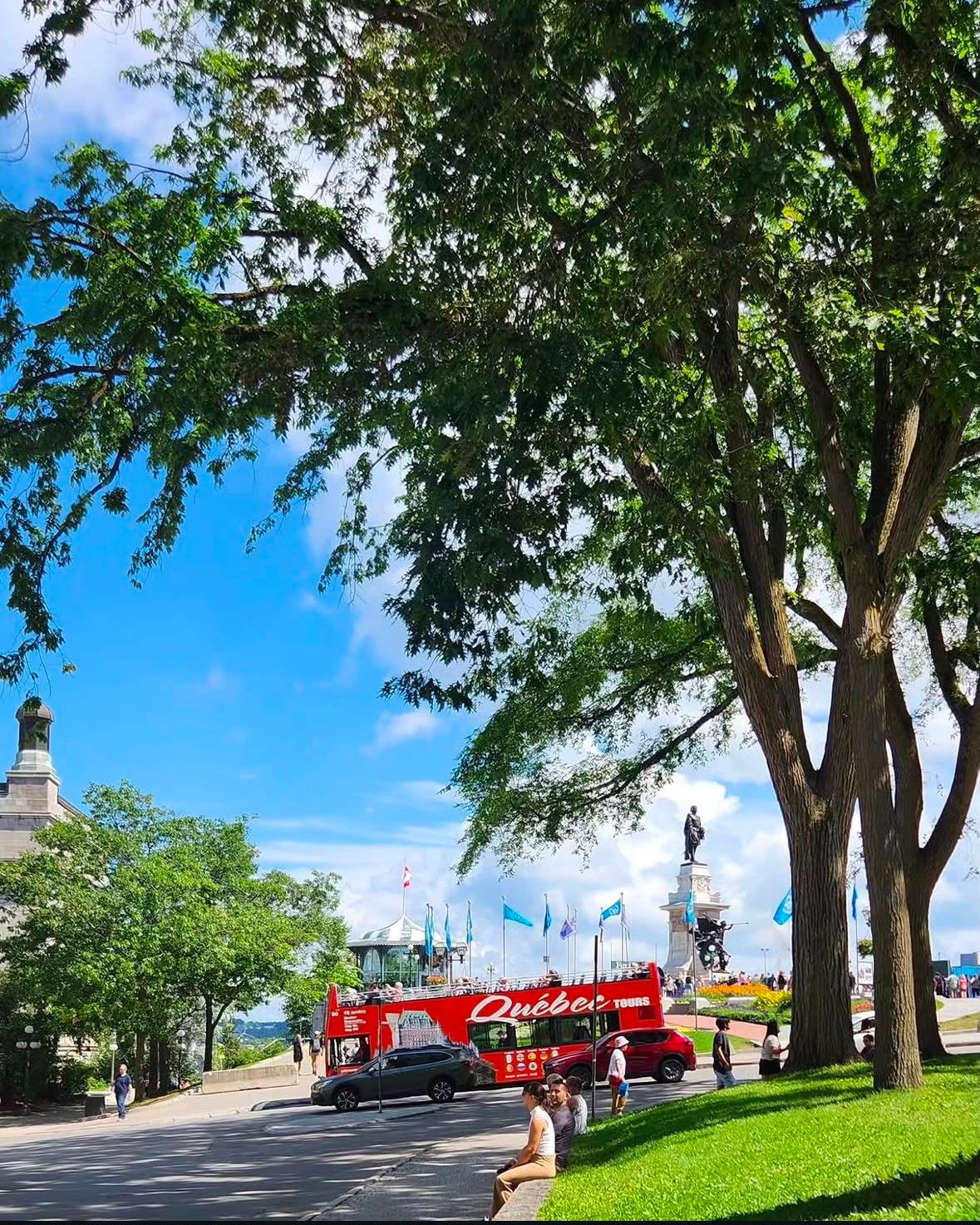 Red Quebec City tour bus driving past a large tree-lined promenade with historic buildings and monuments visible in the background